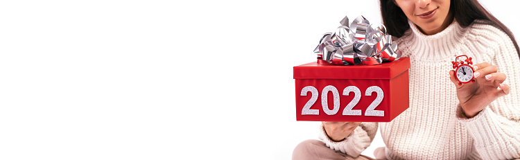 Close up of young woman on a white background holding a red gift box with the numbers 2022 and red alarm clock.The clock shows midnight.Concept of the New year 2022.Selective focus,copy space.