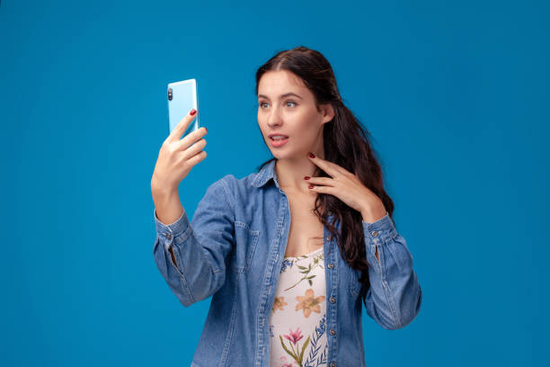 istock Young brunette woman is posing with a smartphone standing on blue background 1355228455