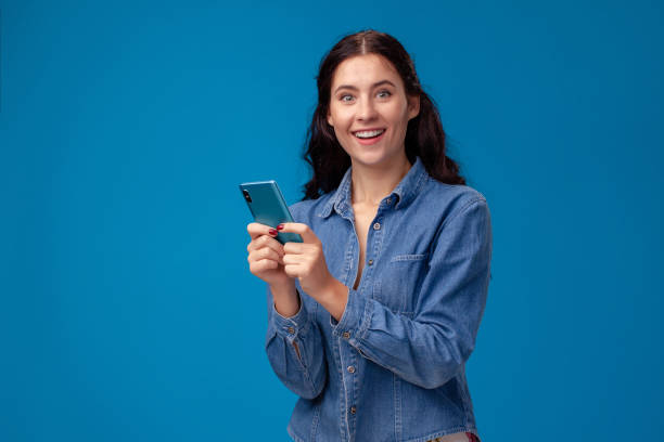 Young brunette woman is posing with a smartphone standing on blue background stock photo