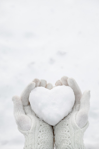 A snow heart in the hands wearing knitted gloves against the background of a snow cover.