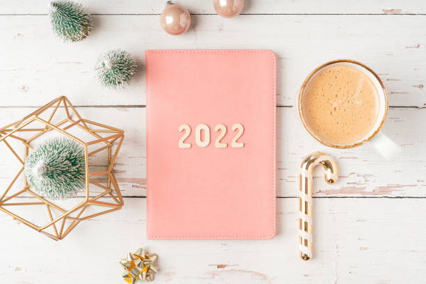 istock Top view Desktop Christmas pink notepad with 2022 letters text. Flat lay on white wooden table background with planner, cup of coffee, candle, toys, Christmas decoration, notebook and stationery 1355243394