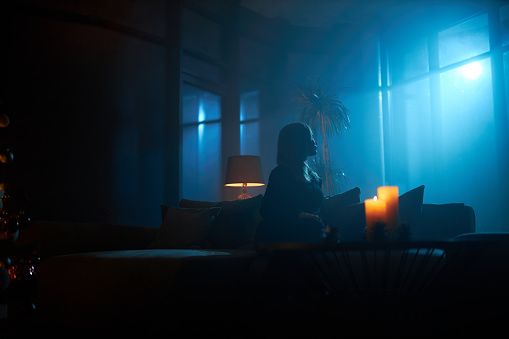 Silhouette of young woman sitting on couch at dark atmosphere and looking on full moon through panoramic windows. Burning candles on table.