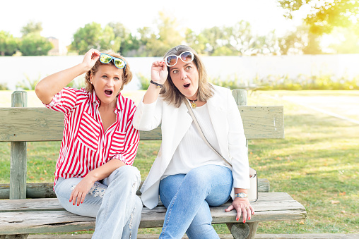 Two middle-aged women sitting on a park bench taking off their glasses to stare. Concept of surprise at some special situation