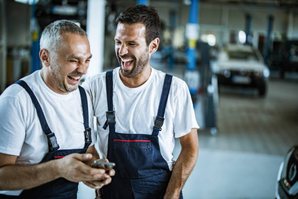 Cheerful auto mechanics using cell phone in a workshop. Cheerful mechanics laughing while using smart phone in auto repair shop. repairperson occupation stock pictures, royalty-free photos & images