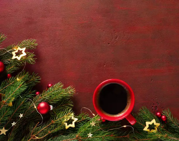 Red cup of coffee and fir branch with Christmas decorations on the wooden board painted in dark-red with copy space for text. Flat lay.