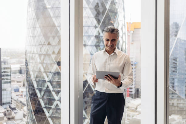London executive using digital tablet in modern office Front view of smiling grey-haired businessman in early 40s standing next to full length window with view of landmark architecture while using wireless device. financial districts stock pictures, royalty-free photos & images
