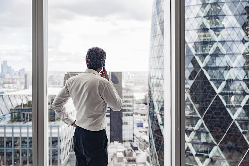 Successful executive in early 40s standing with hand on hip and conversing with client while looking through window at London’s financial district.