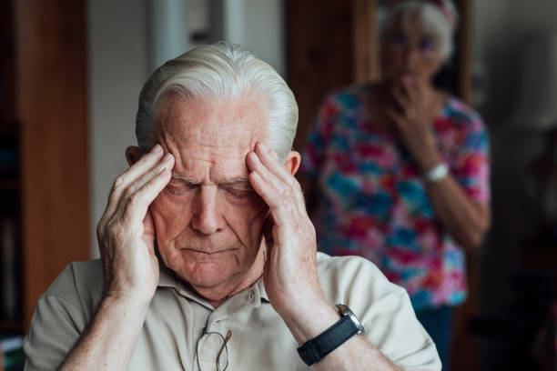 Worried about Him Senior man who has a chronic illness/Alzheimer's disease sitting in a living room at his home in the North East of England. He is holding his forehead with a negative expression, experiencing anxiety/pain. His wife is out of focus behind him, looking at him in worry. dementia stock pictures, royalty-free photos & images