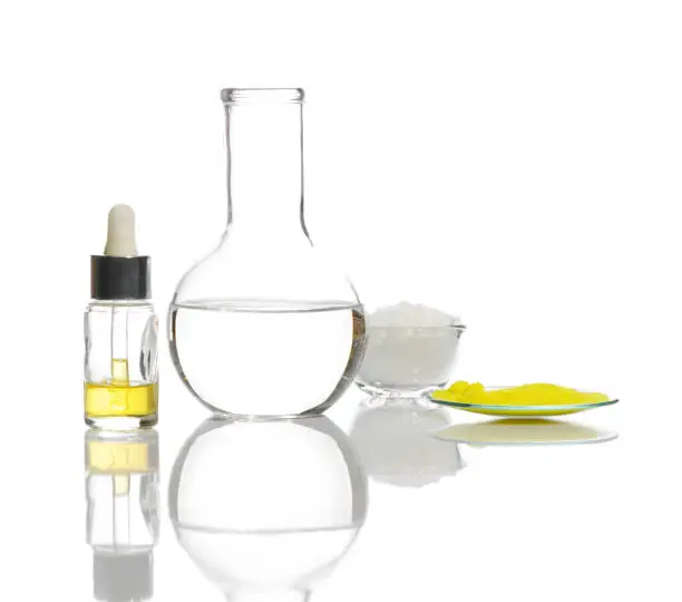 Photo of Closeup chemical ingredient on white laboratory table. Yellow cosmetic liquid in Dropper Bottle, Crystal clear liquid in Flat Bottom Flask, White flake chemical and Potassium Chromate powder.
