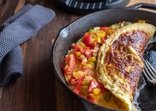 Delicious and healthy omelet with mediterranean vegetables such as tomatoes, bell peppers, chives, zucchini served in a rustic cast iron pan on wooden table.