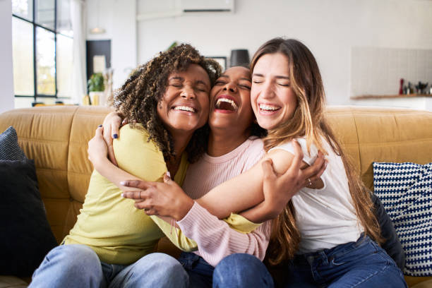 drei glückliche freunde umarmen sich lächelnd. lustige frauen feiern zusammen auf dem wohnzimmersofa - joy of living stock-fotos und bilder