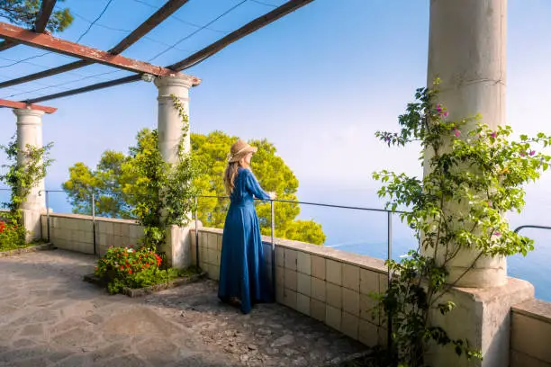A young, blonde girl dressed in blue looking at the sea in Capri Island from the patio of an ancient, historic villa.