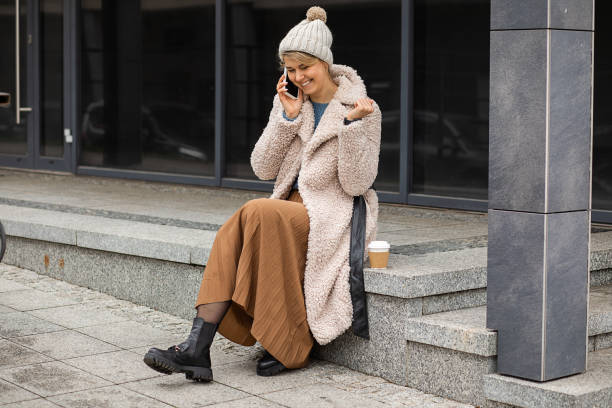 Young cute woman with coffee in disposable cup, using mobile phone, sitting smiling speaking on staircase on european street, wearing fur faux coat and hat, street style, modern life