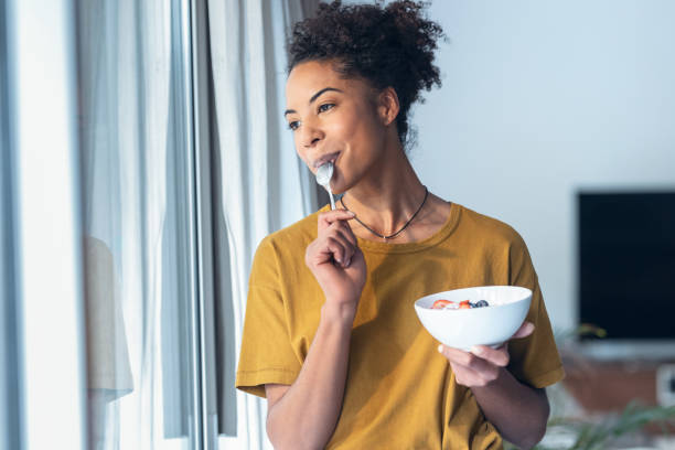 hermosa mujer madura comiendo cereales y frutas mientras está de pie junto a la ventana en casa. - comer fotografías e imágenes de stock