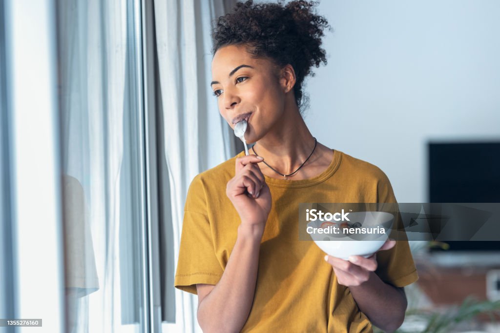 Beautiful mature woman eating cereals and fruits while standing next to the window at home. Shot of beautiful mature woman eating cereals and fruits while standing next to the window at home. Eating Stock Photo