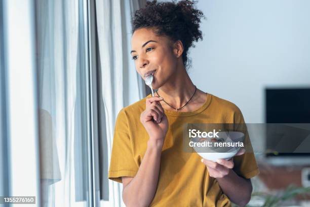 Beautiful Mature Woman Eating Cereals And Fruits While Standing Next To The Window At Home Stock Photo - Download Image Now