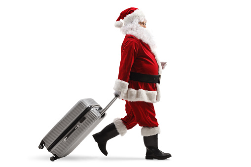 Full length profile shot of santa claus walking and pulling a suitcase isolated on white background