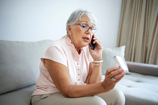 Photo of senior woman at home preparing to drink medical pill, while holding a medication in hands and talking on the phone with doctor. Close up of senior woman consulting with a doctor on his phone.