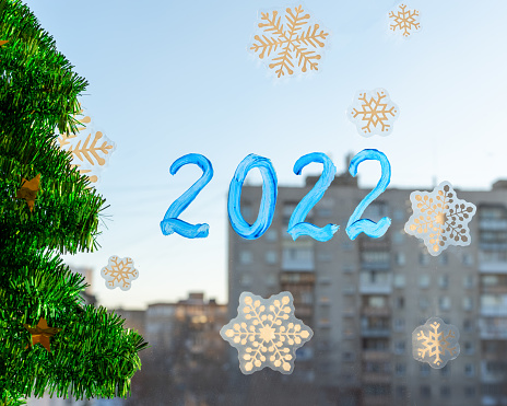 Drawn numbers of the year 2022 on a window pane decorated with stickers with snowflakes and a shiny green artificial Christmas tree. Christmas decoration of the windows of the house.