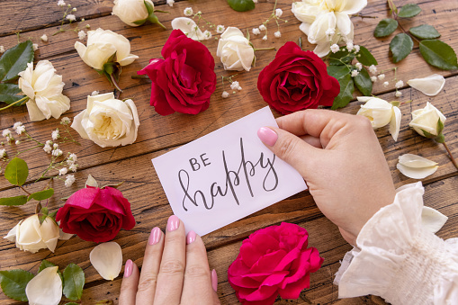 Hands with handwritten card BE HAPPY surrounded by red and cream roses close up on a wooden table. Femminine romantic declaration of love near flowers, Valentines day concept
