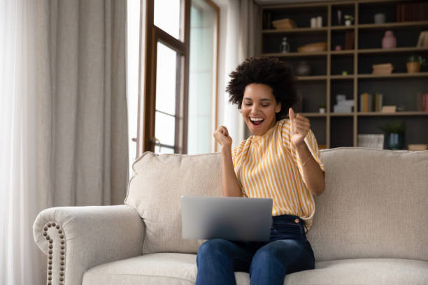 Excited cheerful young Black woman using laptop computer Excited cheerful young Black woman using laptop computer on sofa at home, getting good news, feeling joy, dancing with hands, singing, laughing, making winner gesture, happy to win prize incentive stock pictures, royalty-free photos & images