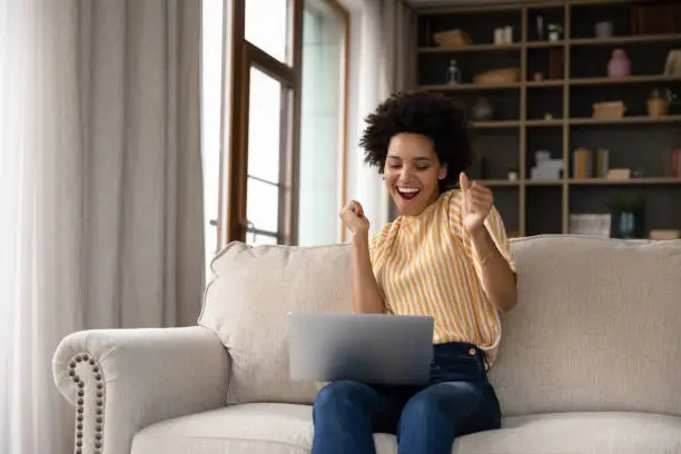 Excited cheerful young Black woman using laptop computer on sofa at home, getting good news, feeling joy, dancing with hands, singing, laughing, making winner gesture, happy to win prize