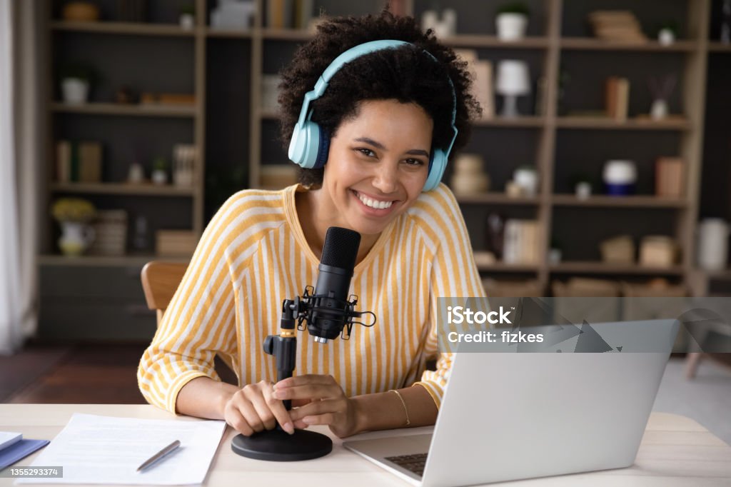 Happy young Black woman in headphones and professional microphone recording Happy cheerful young Black woman in headphones and professional microphone recording audio podcast for radio, live channel on internet, presenting news on air. Newscaster, blogger head shot portrait Podcasting Stock Photo