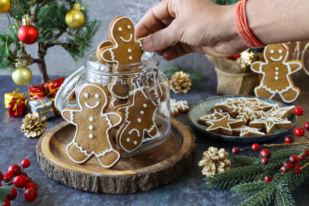 istock Image of unrecognisable person selecting a gingerbread man biscuit, decorated with white royal icing from glass cookie jar of gingerbread people, mini Christmas tree, red and gold baubles, grey background, home baking 1355295792