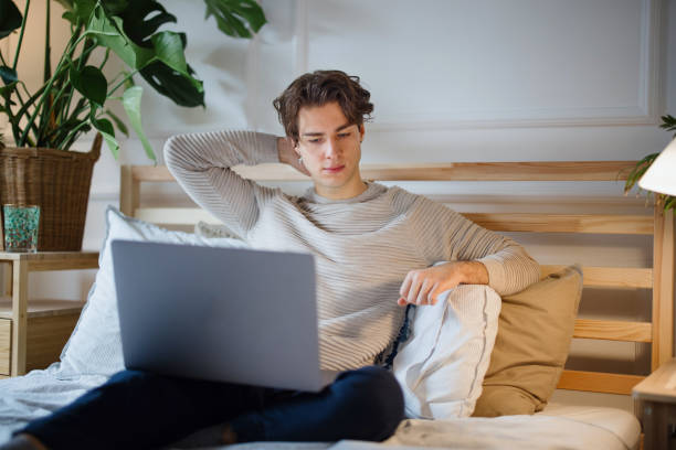 joven relajándose en casa y usando la computadora portátil para navegar por internet - working bed smart phone bedroom fotografías e imágenes de stock