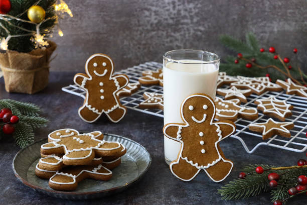 istock Image of gingerbread man biscuit, decorated with white royal icing leaning on drinking glass of milk besides cooling rack and plate of cookies, spruce needles and red berries, grey background, home baking 1355298681