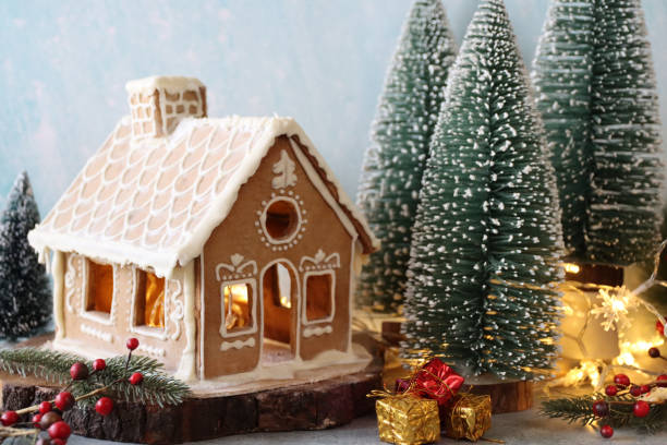Close-up image of homemade, gingerbread house decorated with white royal icing displayed in snowy conifer forest scene, foil wrapped presents, model fir trees, icing sugar snow, snowy blue background stock photo