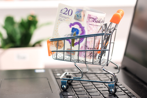 20 Britains pounds placed in supermarket shopping cart on keyboard of laptop computer illustrating online shopping with cash and shipping products to consumers.