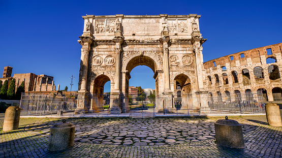 A wide angle view of the stunning Arch of Constantine in the imperial heart of the Eternal City. The largest triumphal arch in the ancient world celebrates the victory of Emperor Constantine over the usurper Maxentius in the battle of Ponte Milvio (Milvian Bridge), which took place in 312 AD near Rome, along the Tiber river. With its 21 meters high, 25.9 meters wide and 7.9 meters deep, the Arch of Constantine is a compendium of Roman sculpture and history, with depictions and bas-reliefs concerning the gestures of the emperors Hadrian, Trajan and Marcus Aurelius. Built in masonry and marble in the core of the Roman Forum, between the imperial hill of the Palatine and the Celian hill, this sumptuous Roman monument is placed diagonally to the Flavian Amphitheater or Colosseum (right in the photo). The Roman Forums, one of the largest archaeological areas in the world, represented the political, legal, religious and economic center of the Ancient Rome, as well as the nerve center of the entire Roman civilization. In 1980 the historic center of Rome was declared a World Heritage Site by Unesco. Super wide angle and high definition image in 16:9 format.