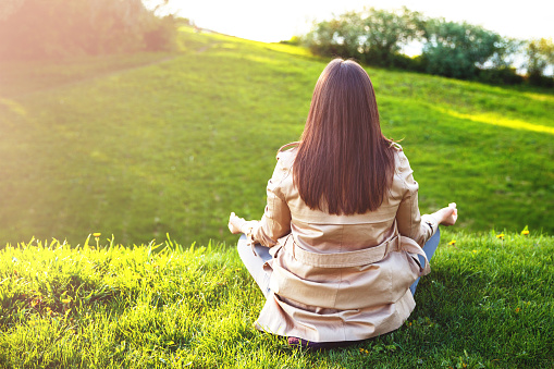 A young girl meditates sitting on the green grass in nature. She has beautiful long brown hair. A woman practices yoga and meditation sitting in a lotus position on the shore of a pond.