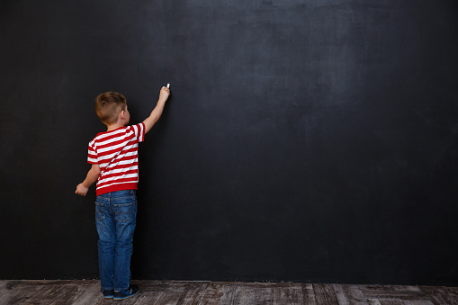 Back view of cute little kid boy writing with chalk on the backboard in school class