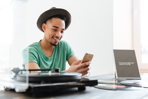 Young African man in t-shirt and hat sitting by the table with record-player and records while using smartphone
