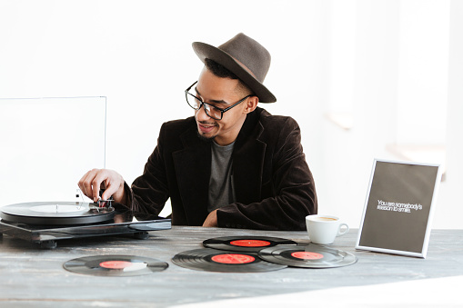 African man in hat, suit and eyeglasses sitting by the table and using record-player