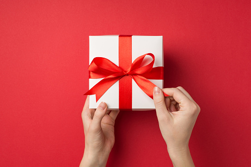 First person top view photo of woman's hands unpacking big white gift box with red satin ribbon bow on isolated red background