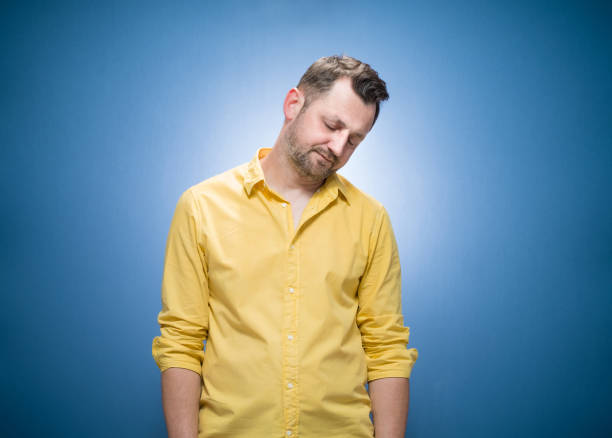 Sleepy guy standing and wearing sleep mask over blue background, dresses in yellow shirt. Exhausted young man. Drowsy expression stock photo
