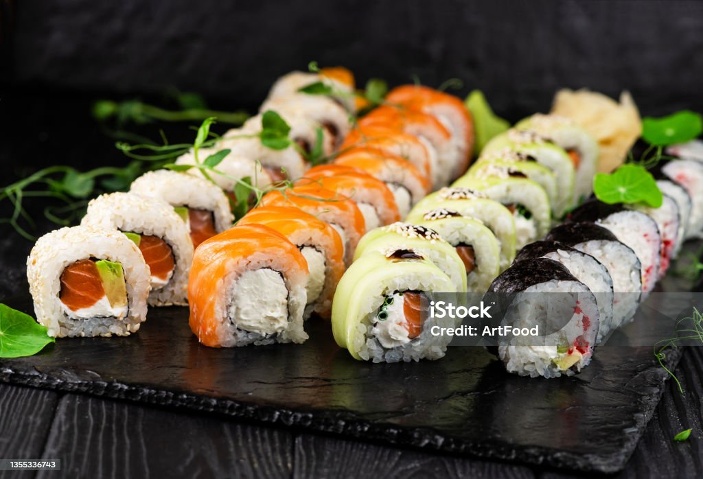 Set of sushi roll pieces on black slate stone plate with sprouts of fresh herbs. Sushi pieces Assortment Set of Japanese sushi roll pieces served on black slate stone plate on wooden background with sprouts of fresh herbs. Assortment of Sushi pieces with salmon, avocado and cream cheese Sushi Stock Photo