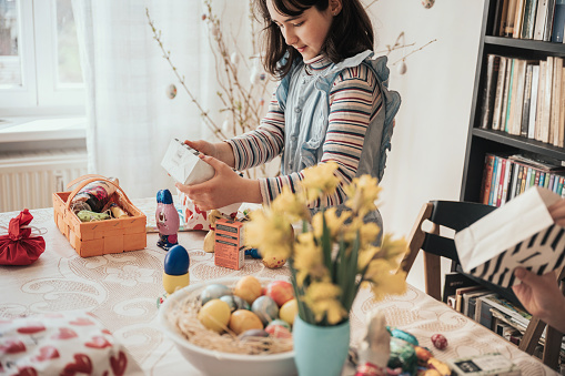 little girl looking at gift box in hands at easter table with basket filled with colorful easter eggs in living room