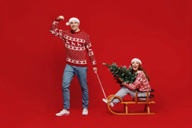 Full size young couple friends two man woman 20s in sweater hat hold fir-tree ride sled isolated on plain red background studio portrait. Happy New Year 2022 celebration merry ho x-mas holiday concept