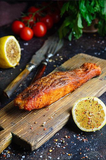 Grilled salmon steak served on wooden board with ingredients and spices on dark background. Lemon, tomatoes and green herbs near dish.