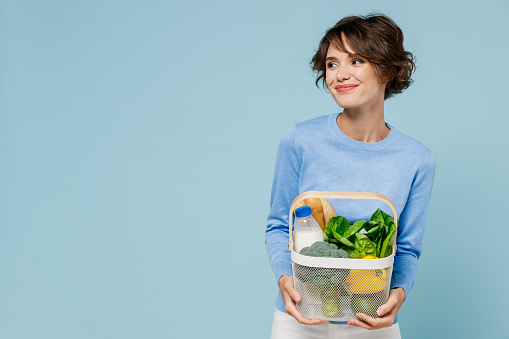 Young smiling woman in casual sweater look camera hold white busket with begetables look aside on copy space ingredients isolated on plain pastel light blue background. People lifestyle food concept