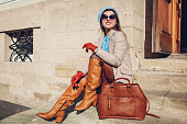 Portrait of woman wearing stylish orange boots coat beret sitting on stairs by handbag outdoors. Fall female fashion.