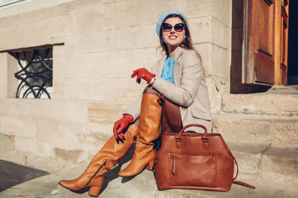 Portrait of woman wearing stylish orange boots coat beret sitting on stairs by handbag outdoors. Fall female fashionable accesories. Vintage outfit
