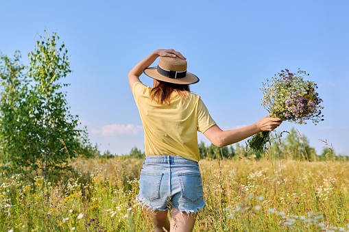 Summer season, young woman in straw hat running in meadow with bouquet of wild flowers, back view. Summer, nature, summertime, beauty, woman concept.