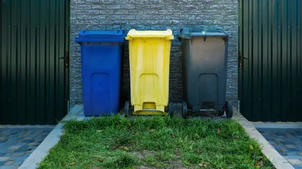 Blue yellow and grey plastic trash bins stands in row on green grass on backyard of town street. Pollution and environmental care concept.