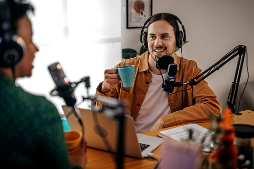 Smiling young man with headphones drinking coffee and recording podcast over microphone at recording studio with women