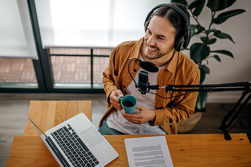 Top view of young handsome smiling man recording podcast with headphones, microphone, laptop and paper drinking coffee in recording studio